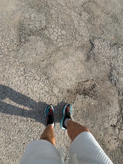 A first-person, downward-angle photograph depicts the lower legs and feet of an individual standing on a textured, light grey asphalt or compacted gravel surface in Kermen, Bulgaria. The subject wears black athletic shoes featuring prominent turquoise, red, and orange/coral lacing or design patterns, along with grey socks and white, textured shorts. Distinct shadows from the individual's upper body and limbs are cast to the left side of the frame, indicating direct sunlight from the right. The ground shows irregular patching, small gravel, and variations in surface integrity. No other individuals or objects are visible.