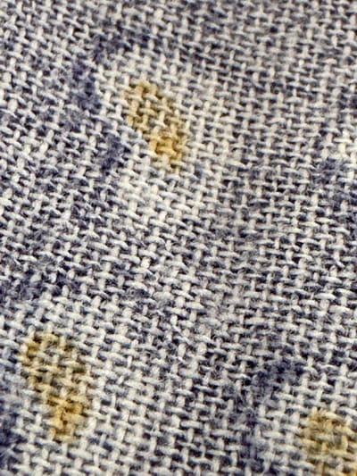 This is a close-up, macro photograph of a coarse, woven fabric with a blue and white checkered pattern. Two prominent yellow-brown stains are visible on the fabric. The image is a detailed examination of the fabric's texture and the stains, without any contextual information about its location in Kyzyl-Kyya, Kyrgyzstan, or any other environmental or human elements.