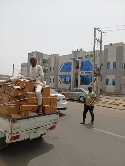 The image displays an urban street scene in Kofar Mazugal, Nigeria, under a bright, clear sky. A white pickup truck occupies the left foreground, its bed heavily loaded with numerous brown cardboard boxes secured by white ropes. A man, wearing a light-colored traditional garment and cap, is seated directly on top of these stacked boxes in the truck bed. To the right of the truck, on the dark gray paved road, a second man stands, dressed in an olive green t-shirt and dark trousers, facing slightly left. The road exhibits a white dashed line. In the background, a multi-story building with a light-colored facade and prominent blue-tinted windows is visible. Signage on this building reads "THE NEW AKIJA" and "BIOHA GLOBAL LIMITED," with "ASHEHA TRAVELS & TOUR" also present. A tall utility pole with an electrical transformer stands in the mid-ground, to the right of the building. Two sedans, one white and one gold, are partially visible in the mid-ground behind the truck and the standing man.