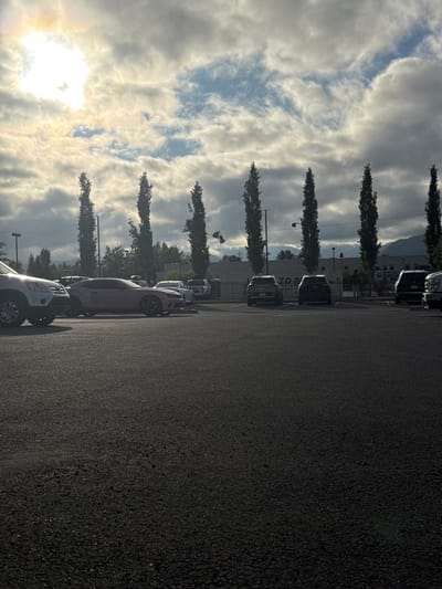 This media file shows a parking lot scene with a row of cars parked along an asphalt surface. The image is oriented vertically. Tall, slender trees with dark foliage are lined up along the left side of the frame, casting shadows on the parking lot. In the background, there are buildings, one of which has visible signage. The sky is filled with dramatic, textured clouds, with the sun shining brightly through them, creating a halo effect and illuminating the scene with a warm, golden light. The time of day appears to be either early morning or late afternoon, given the low angle of the sun and the quality of the light. The weather seems to be partly cloudy, with a mix of sun and clouds.

Several cars are visible, parked one behind another. The closest car in the foreground is a white pickup truck. Behind it, a purple sports car is prominently featured. Further down the row, other vehicles, mostly dark-colored SUVs and sedans, are parked. The overall mood is somewhat serene, with the dramatic sky and the quiet parking lot.

Visible text includes "TOYOTA" on one of the buildings in the background. The scene appears to be a commercial area, possibly a dealership or a business park. The location context provided is Anchorage, United States of America.