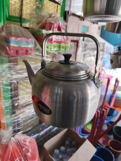 The image captures a retail scene in Dayahbaro, Indonesia, specifically an area displaying household goods. A silver-toned aluminum kettle with a black lid knob and an arched metal handle is centrally positioned and in sharp focus. A black and red product label, bearing the text "aawa" and "aluminum group," is affixed to its side. The background, largely out of focus, reveals a dense assortment of merchandise. To the left, several brightly colored plastic items, resembling baskets or containers with patterns of pink, green, blue, and red, are individually encased in clear plastic wrap and appear to be hanging on a display structure with green elements. To the right, red-handled cleaning tools, such as brooms or mops, are vertically arranged, alongside various colored plastic buckets in shades of blue, brown, and black. A smaller, similar aluminum kettle is partially visible hanging in the upper right. The lower foreground includes an open cardboard box containing numerous small plastic bottles, likely filled with water, and a transparent plastic bag holding orange plastic items. No individuals are present, and no actions or interactions are depicted.