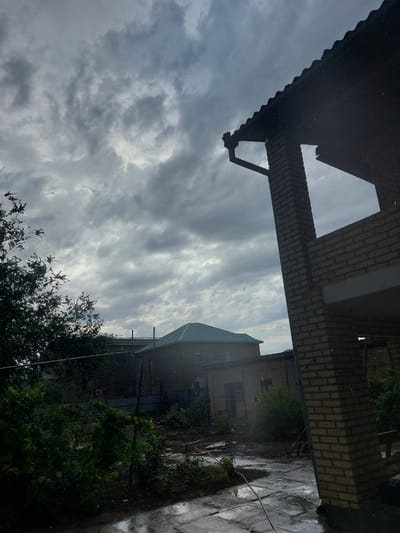 The image depicts an overcast sky with dramatic cloud formations, suggesting a potentially stormy or moody atmosphere. In the foreground, a dark, corrugated roof structure casts a shadow, with a gutter visible. Beyond this, a brick building with lighter-colored bricks is partially visible, featuring a prominent triangular architectural element. Further in the background, another building with a teal-colored roof is seen, with windows and what appears to be a downspout. The ground area, likely a patio or walkway, is wet and reflects the sky, indicating recent or ongoing rain. Green foliage and trees are present, adding to the outdoor setting. The overall scene appears to be a residential or semi-commercial area in Kyzylorda, Kazakhstan, possibly captured during daylight hours due to the sky's illumination, but the heavy cloud cover obscures direct sunlight. There is no visible text in the image.