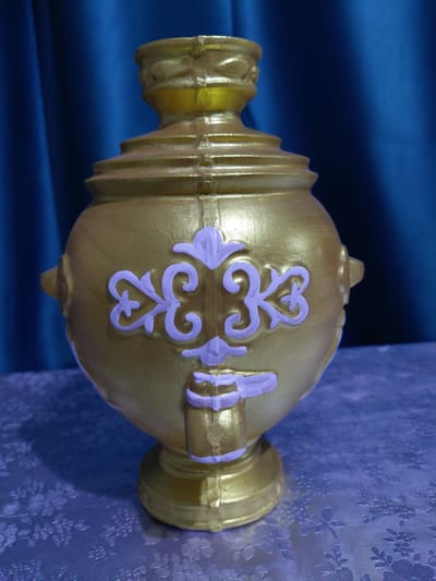 This is a close-up, rotated, horizontal shot of a golden vase with decorative purple accents. The vase has a bulbous body with a narrow neck and a flared base. It is adorned with ornate, scroll-like designs in a lighter purple color, creating a striking contrast against the metallic gold. The vase appears to be made of plastic or a similar molded material.
The background is a blend of textures and colors. On the left, a dark blue fabric with a smooth, draped surface is visible. On the right, a textured, patterned fabric in a lighter blue or violet hue, possibly with a floral motif, provides a backdrop. The lighting suggests an indoor setting, casting subtle shadows and highlighting the reflective qualities of the vase.
There is no visible text in the image. The image does not depict any people or show any specific event or activity. The focus is solely on the object and its immediate surroundings, emphasizing its decorative and possibly cultural significance. The scene is static and presented in a studio-like manner. The location context of "Ертіс ауылдық округі, Kazakhstan" suggests this object might be part of a local display or cultural representation within that region.