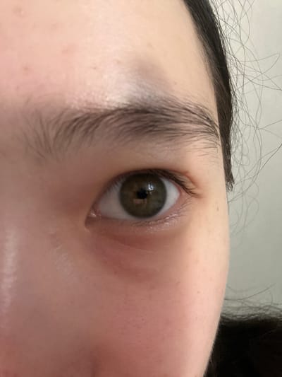 A close-up, eye-level, medium shot focuses on a person's right eye. The eye is open, with a brown iris and dark pupil. The skin around the eye is fair, with some slight redness and visible pores. The eyebrow above the eye is dark and appears naturally shaped. Strands of dark hair are visible on the right side of the frame, and a light-colored wall serves as the background. The image does not contain any information that would allow for the determination of its location in Nookat, Kyrgyzstan, or any other geographical area.