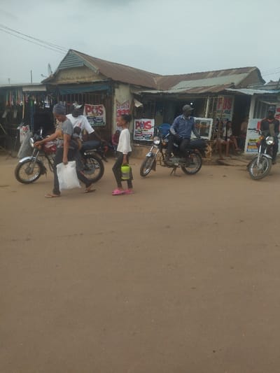 The scene is a street in Junction, Nigeria (8.958, 7.551). There are four people, two women and two men, standing in the street. The women are walking, while the men are standing. There are also two motorcycles and a building with a shop in the background. The shop has a sign that says "POS" on it. There are various items for sale outside the shop, including brooms, plastic chairs, and other assorted items. The road is unpaved and dusty.  The sky is overcast.  The image was taken during the day.