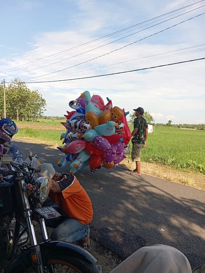 The image depicts an outdoor scene in the Waung area of Indonesia. A person wearing a green camouflage shirt and khaki shorts is standing next to a large cluster of colorful, cartoon-themed balloons that are tied together, appearing to be for sale. An adult in an orange shirt is crouched near a motorcycle, possibly working on it. Another individual wearing a blue baseball cap with cartoon characters is partially visible near the motorcycle. The environment includes a paved road, a green field, and a blue sky with white clouds. Utility lines are visible in the sky. The scene is in daylight.