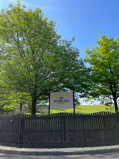 The image shows a wooden fence with a sign that reads "Woburn Golf Club" in front of a golf course. Two large green trees stand in the foreground, with a smaller, thinner tree in the background. There are people in the background on the golf course. The sky is blue. The scene is located near Woburn Golf Club, in Woburn, Bedfordshire, UK.  The coordinates (latitude: 51.991, longitude: -0.663) are consistent with the location of Woburn Golf Club.