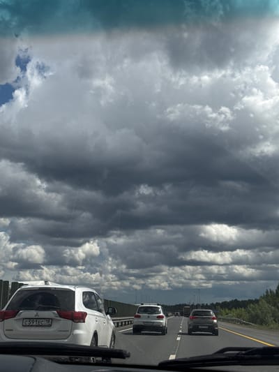 The image depicts a view from inside a vehicle traveling on a highway. Several cars are visible on the road ahead. The vehicle closest to the camera is a white Mitsubishi Outlander, with a license plate number C 591 EC 18. Behind the Outlander are other cars, including a grey sedan. The road is lined with trees and a guardrail. Overlooking the highway is a cloudy sky with varying shades of gray. The city of Nagornaya, Russia, is the location in which this image was taken.