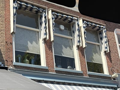 The image shows the upper facade of a brick building in Steenwijk, Netherlands, featuring three windows with striped awnings and internal blinds. The windows are framed by white decorative elements. The building's brickwork is visible, and a dark, curved object occupies the upper right portion of the image, partially obscuring the sky. Below the windows, a grey awning with blue and white stripes is visible. The scene appears to be an exterior view of a shop or residential building.