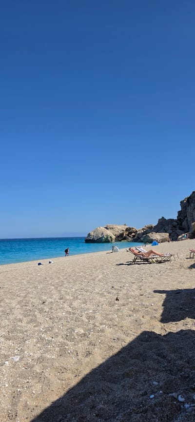 This vertical image depicts a sunny beach scene within the Municipal Unit of Lefkada, Greece. The upper half of the frame is dominated by a clear, azure blue sky. Below, a vibrant turquoise sea meets a wide expanse of light brown and tan pebbly sand. In the mid-ground, a person wearing red shorts and a dark top or backpack is walking near the water's edge, heading towards the right. Close by, two small dark items rest on the sand. Further to the right, a blonde individual dressed in a dark swimsuit reclines on a light-colored sun lounger. Behind the sun lounger, a small, gray, dome-shaped beach tent is visible. The background features large, light-colored rock formations and darker cliffs extending along the coastline and into the sea. Several indistinct figures are present near the water and rocky areas in the distance. A prominent, elongated dark shadow cuts diagonally across the sandy foreground from the lower right corner.