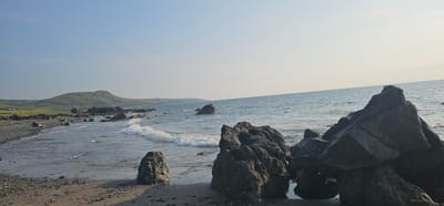 The image shows a scenic view of a rocky beach with the ocean in the background. The foreground features large, dark, angular rocks, some of which are partially submerged in shallow water. A smaller, rounded rock sits on the wet sand further back. Waves are gently breaking on the shore, creating white foam. The beach itself is a mix of sand and small pebbles. In the mid-ground, a line of dark rocks extends into the water. Beyond the water, a green, rolling hill dominates the landscape, with a few scattered buildings visible on its lower slopes. The sky is clear and bright, with a soft gradient from a pale blue at the top to a lighter, almost white hue near the horizon, suggesting it's either early morning or late afternoon. The scene appears to be a natural coastal environment in Girvan, United Kingdom.
