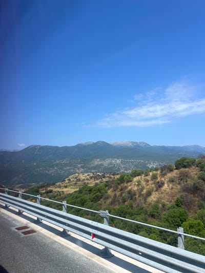 The image depicts a scenic view of a mountainous landscape under a clear blue sky, likely taken from a moving vehicle on a road in or near Tsangaropoulo, Greece. In the foreground, a grey metal guardrail with cables runs along the edge of an asphalt road. The road surface is visible in the lower left corner. Beyond the guardrail, a dry, grassy hillside dotted with low green bushes slopes upwards. Further in the distance, layers of green, tree-covered mountains form a panoramic backdrop, their peaks tinged with a hazy grey. A few wispy clouds are present in the sky. The overall impression is one of a bright, sunny day in a rural, mountainous region.