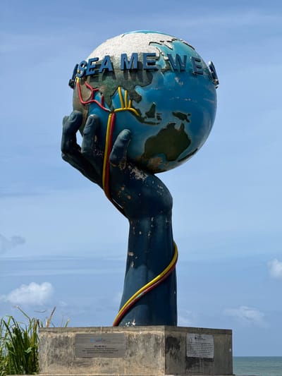 This is a full-shot, eye-level, outdoor photograph of a large monument in Dondra, Sri Lanka. The monument, which stands on a concrete base, depicts a dark blue, open hand holding up a globe of Earth. The globe has the words "SEA ME WE 5" written in blue lettering around its circumference. Red and yellow cables wrap around the hand and the globe, connecting to what appears to be a representation of submarine cable infrastructure. The monument is set against a bright blue sky with scattered white clouds. The background shows a body of water, likely the ocean, and some green foliage on the left side of the frame. A plaque with text is affixed to the front of the concrete base.