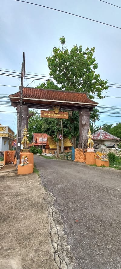 The image depicts a street scene in Ban Thung, Thailand. A large, ornate, wooden archway with a red-tiled roof stands over the road. A sign with Thai script hangs beneath the arch. Trees grow around the arch, and power lines are visible overhead. Concrete posts decorated with golden figures line the roadside. Buildings with traditional Thai architecture are visible beyond the arch. The road is paved with asphalt, showing signs of wear and cracking.