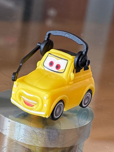 A yellow toy car with headphones is displayed on a clear, circular platform. The car has large red eyes with black pupils and a wide smile with red lips. The headphones are black and rest on top of the car. The car is positioned slightly to the left of the center of the image. The background is blurred and appears to be a wooden surface. The scene is lit from above, casting a slight shadow on the platform. There is no indication of the car's location in Toruń, Poland.