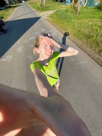 A person with blonde hair, wearing a bright green t-shirt and colorful shorts, is lying on asphalt. They have tattoos on their left arm and are smiling with their eyes closed, hands resting on their head. The ground around them is marked with chalk drawings, including what appear to be butterflies and abstract shapes. The scene is outdoors, with a grassy area and trees visible to the left. A shadow is cast on the asphalt, indicating it is daytime, likely sunny. The context provided places the location in Сям-Можга, Russia. Visible text on the t-shirt is partially obscured but appears to be in English, possibly related to an event or organization. The overall mood conveyed by the image is one of joy and relaxation on a sunny day.