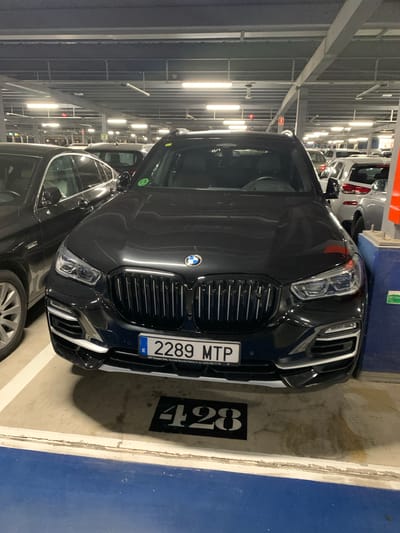 The image shows the front of a black BMW SUV parked in an indoor parking garage. The vehicle has its headlights on and its license plate is visible, reading "2289 MTP". On the ground in front of the car, the number "428" is painted in a black square. The parking garage appears to be well-lit with fluorescent lights. The surrounding area shows other vehicles parked, including a dark-colored sedan to the left and other cars in the background. The image is taken from a slightly low angle, looking up at the front of the BMW. The location is identified as Manises, Spain, implying this is a parking facility in that city.