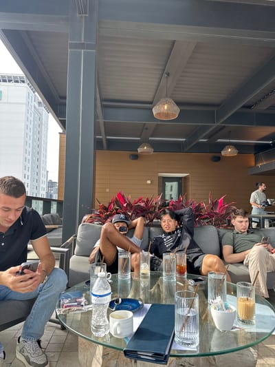 The scene depicts a group of four people sitting in an outdoor seating area, likely a rooftop bar or lounge. The location appears to be Tampa, Florida, suggested by the distant view of buildings resembling the city's skyline. The area is shaded by an overhead structure of metal beams and wicker lampshades.

A round glass table stands in front of the group, covered with several empty drinking glasses, a water bottle, a sugar holder, and a blue folder. The group is sitting on a grey upholstered couch.

One man, clad in a black shirt and blue jeans, is looking at his phone. Two individuals are casually lying back on the couch: one with a black shirt, and the other with a green shirt. A fourth man is sitting farther away.

The setting includes red tropical plants between the couches and the exterior wall.