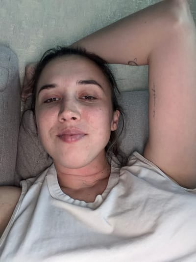 A person is lying on their side on a sofa, their head resting on their arm. They are wearing a light-colored t-shirt and have long, dark hair. The person's face is turned towards the camera, and they appear to be looking directly at it. Their eyes are open, and their mouth is slightly parted. There is a tattoo on their upper arm, with some visible text. The setting appears to be indoors, with soft lighting, suggesting it might be daytime. The overall mood conveyed is relaxed and intimate. The visible text on the arm reads "no regrets".