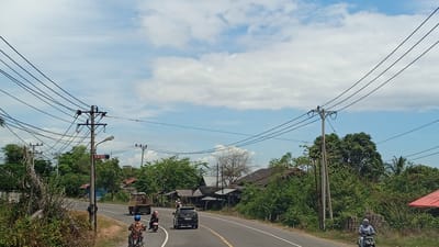 The image shows a roadside scene in Lhokdagang, Indonesia, under a partly cloudy sky. A two-lane asphalt road curves through a rural area with lush green vegetation and scattered houses. Several utility poles with numerous wires are prominent along the roadside. In the foreground, a person on a motorcycle wearing a blue helmet rides in the right lane. Further down the road, another motorcycle is being ridden by a person wearing a hijab. A black car is also traveling in the right lane. On the left side of the road, a large truck is parked or moving slowly. In the background, traditional Indonesian houses with reddish-brown roofs are visible amidst dense trees and foliage. A sign on a utility pole reads "SPBU," indicating a gas station nearby. The overall impression is of a typical day in a developing region of Indonesia, with local transportation and infrastructure.