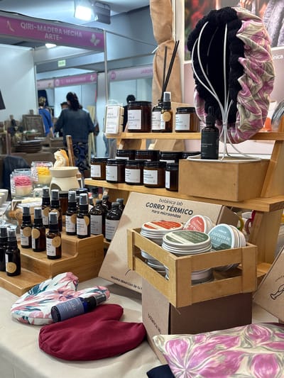 The image shows a stall at an artisan market. In the foreground, multiple shelves display bottles, jars, and containers of what appear to be beauty or wellness products. The products are arranged in a tiered manner, making efficient use of the space. The shelving and displays are made of wood.
The bottles and jars are labeled with text in Spanish, including product names and descriptions like "GORRO TÉRMICO Para microondas" and "UNGUENTO DE UÑA DE GATO." The products are from a company called "botánica lab."
Behind the displays, a woman with dark hair, wearing a denim jacket and a mask, is seen browsing.
The background reveals elements of the market setting, with a sign that reads "QIRI-MADERA HECHA ARTE-" along with a drawing of a foot and shoe, indicating that some of the vendors are selling wooden artifacts.
Based on the text "QIRI-MADERA HECHA ARTE-", the location is in the city of Chorrillos, Peru.