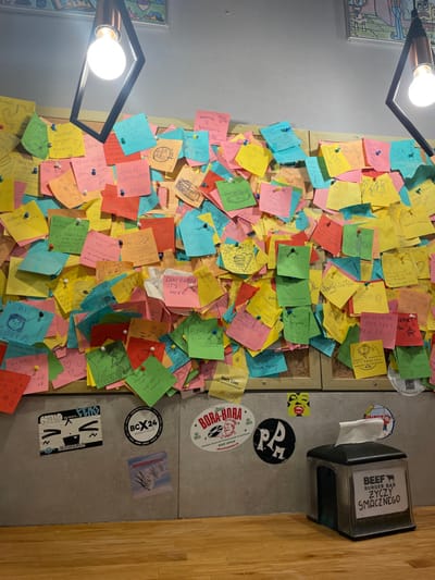 The image depicts an indoor scene from what appears to be a casual eatery or burger bar, located in Kraków, Poland. The most prominent feature is a wall densely covered with hundreds of colorful sticky notes (Post-it notes) of various hues, including yellow, pink, blue, green, orange, and red. Each note is held in place by a small pushpin, forming a vibrant, mosaic-like display. Many of these notes bear handwritten messages, doodles, and simple drawings, indicating a space where patrons leave personal remarks or reviews.
To the right, a section of grey wall, possibly tiled or concrete, is visible, adorned with several stickers, including "BORA-BORA," "BCX24," and a black circular one with "PA" in white. A black plastic napkin dispenser is mounted on this wall section, clearly displaying the text "BEEF BURGER BAR ZYCZY SMACZNEGO," which translates from Polish to "BEEF BURGER BAR WISHES YOU A TASTY MEAL." Part of a wooden floor is visible on the far right.
Artificial lighting from two modern fixtures with exposed bulbs illuminates the area, suggesting it is either evening or an interior space relying solely on artificial light. The colorful and personalized messages on the wall create a sense of community and customer engagement. Other decipherable texts on the sticky notes include "DANI PINEDA IT'S HERE," "Nett hier." (German for "Nice here."), "GAETA," "KOTEC," "TYDANCERS," and "GPERORE."