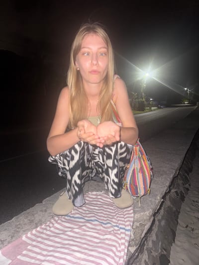 A young blonde woman with blue eyes and light skin is crouching at night on a low concrete wall in Mare Anglaise, Seychelles. She is wearing a light green tank top, black and white patterned pants, and beige slip-on shoes. She has her hands cupped together in front of her. A colorful crocheted bag with fringe is slung over her right shoulder. In front of her, on the wall, is a pink and white striped towel. The background shows a dark asphalt road with streetlights illuminating the scene, and some vegetation is visible. The lighting suggests it is nighttime, and the overall setting appears to be along a coastal road.