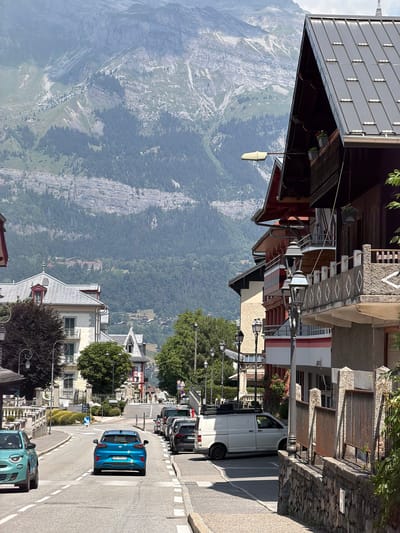 The image depicts a view looking up a street in Saint-Gervais-les-Bains, France, towards a mountain range. The street curves to the left and uphill. On the right side of the street, a multi-story building with a dark metal roof and balconies is prominent. The building has a stone base and detailed decorative elements on its upper balconies, including patterned railings. Several street lamps with ornate designs line the sidewalk. A white van is parked on the right side of the street, and several other vehicles are visible further up the road.
On the left side of the street, there are other buildings, including a lighter-colored building with a red-roofed dormer window and balconies. A teal-colored car is driving away from the viewer on the left side of the street, and a blue car is driving away from the viewer in the center of the street. Trees and vegetation are present along the street and on the slopes of the mountains in the background. The mountains themselves are large and rugged, with visible rock formations and patches of trees. The sky is bright and appears to be partly cloudy. The overall scene suggests a daytime view in a mountainous town.