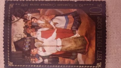 The image is a close-up of a postage stamp. The stamp depicts a painting of a group of people in traditional clothing. The scene appears to be set in a rural or historical context, with a building and trees visible in the background. The people are interacting with each other, and some are holding objects. The stamp has text in Russian, including "10. В. Карапаева. Северная песня 1970" and "1977 · ПОЧТА СССР". The stamp is dark blue with a serrated edge.