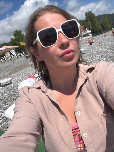 The image shows a woman taking a selfie at a pebble beach in Tsandrypsh, Abkhazia. She is wearing oversized white sunglasses and a light brown open-collared shirt. Her lips are slightly parted, and her hair is light brown and slightly damp. Behind her, the beach is covered in grey pebbles. In the background, several people are visible lounging on the beach, and some beach umbrellas and lounge chairs are set up. Trees and a white building with a balcony are also present in the background, along with a clear blue sky with scattered white clouds. The overall scene depicts a sunny day at the seaside resort.