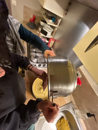 A low-angle shot showcases a kitchen scene. A person in a dark hoodie and long sleeves, with visible wrist accessories, is holding a large metal pot over a round pan filled with yellow batter. The pot is centered in the frame and red-handled. A gas stovetop is visible beneath the pot. To the right, a stainless steel sink is partially visible, with a yellow cloth resting inside. The background includes kitchen cabinets, a soap dispenser, and other miscellaneous items. The scene is dimly lit.