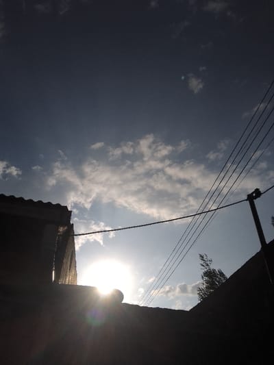 The image depicts an outdoor scene in Njoro ward, Kenya, captured with significant backlighting. The sun is prominently featured as a bright, circular disc with visible lens flare, positioned in the lower-center portion of the frame, suggesting either sunrise or sunset. The sky above is a light blue, partially covered by scattered white cumulus clouds. In the foreground, silhouetted elements dominate the lower half of the image. To the left, the dark silhouette of a building's roofline and upper wall is visible. To the right, a utility pole with at least five parallel overhead power lines extends diagonally across the sky. Below the power lines and pole, a dark, indistinct mass, possibly a distant structure or elevated ground, is present, with a small portion of a tree's canopy visible as a silhouette against the brighter sky. No individuals or specific actions are discernible.