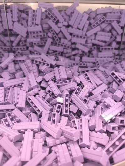 The image shows a bin full of lavender-colored Lego bricks. The bin appears to be overfilled, with the bricks stacked densely and reaching the top of the container. The environment suggests a retail setting, possibly a Lego store. While the exact location in Leidschendam, Netherlands, is not explicitly depicted, it could be a store that sells Lego products within the city. The focus is solely on the mass of Lego bricks within the bin.