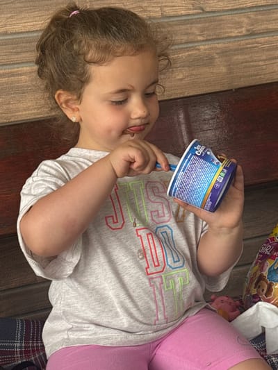 A young Caucasian girl with curly brown hair pulled into a ponytail is seated, holding a small cup of what appears to be ice cream and a blue spoon. She is wearing a light grey t-shirt with colorful lettering that reads "JUST DO IT" and bright pink shorts. Her tongue is slightly sticking out as she appears to be eating the ice cream. She has a small stud earring in her left ear. The background consists of a wooden bench. To the right of the girl, a colorful bag with cartoon characters is partially visible, along with a white tissue and a small pink toy. The lighting suggests an outdoor setting, possibly on a warm day. The image was taken in Rusokastro, Bulgaria.