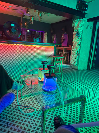 A high-angle, slightly distorted perspective reveals an interior space featuring a hookah, a rotating fan, and decorative elements.

The hookah is positioned on a mirrored surface with a geometric pattern. It consists of a blue-lit container, a vertical stand, and a hose. A person wearing a blue sneaker and dark trousers is visible near the hose's end.

Adjacent to the hookah is a black metal structure with a red and orange lit mesh panel. Behind it, a rotating black fan is situated.

The ceiling and upper walls are decorated with floral elements and lighting fixtures. Pipes run along the ceiling. The walls feature a geometric pattern and green lighting.

The exact location in Drogheda, Ireland, is not evident from the visual information provided.