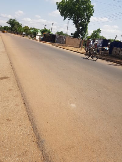 The image depicts a street scene in Palade, Nigeria. A man is seated on a bicycle near some roadside structures. The structures appear to be shops or small businesses, with corrugated metal roofing and brick walls. There are trees and utility poles visible along the roadside. The road itself is paved with asphalt and shows signs of wear and tear. The sky is blue with scattered clouds, suggesting a clear day. The lighting indicates it is daytime, likely late morning or early afternoon. There is no explicit indication of the man's emotional state.

Notable details include the presence of power lines crisscrossing the sky and the style of the roadside businesses. The bicycle is a standard model with two wheels and a frame. The man is wearing casual clothing. The overall impression is of a typical, somewhat busy, urban or semi-urban environment in Nigeria.