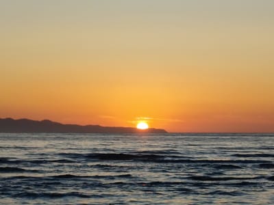 A serene, picturesque sunset unfolds over the Aegean Sea, casting a warm, golden glow across the sky and water. The sun, a brilliant orb, is just beginning to dip below the horizon, its radiant light painting the sky in soft hues of orange, yellow, and a hint of pale blue at the top. In the distance, the silhouetted outline of a mountainous coastline creates a dramatic backdrop against the vibrant sky. The sea itself is calm, with gentle waves creating a rhythmic ripple effect, reflecting the sky's warm colors and darkens as it moves towards the foreground. The scene evokes a sense of peace, tranquility, and natural beauty, characteristic of a Greek island sunset. The time of day is clearly dusk, and the weather appears clear and pleasant. The location is identified as Agios Stefanos, Greece, which is consistent with the coastal landscape and the characteristic light of the Aegean. There is no visible text in the image.