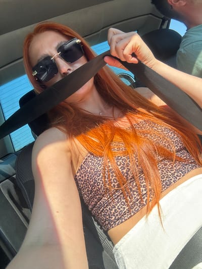 The image depicts two individuals in the interior of a vehicle, likely during the daytime. The foreground is dominated by a person with long, bright red hair, wearing large, square-shaped sunglasses with gold accents. They are seated, and a black seatbelt is prominently visible across their chest. Their attire consists of a leopard-print top.
To the left and slightly behind, another person is partially visible. They are wearing a green t-shirt. Their arm is extended, and their hand is gripping the black seatbelt. The setting appears to be a car interior, with hints of car seats and potentially a window in the background, showing a bright, possibly sunny sky. The lighting suggests it is daytime, with sunlight casting shadows and highlighting certain areas of the subjects and the car interior. The overall mood seems casual and relaxed, possibly indicating a road trip or a drive. No visible text is present in the image. The location context of Medinski Krš, Montenegro, is provided externally and not discernible from the image itself.