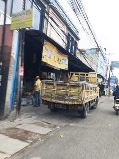 A yellow truck sits in front of a storefront. The building has a yellow banner hanging over the entrance and a sign above it. A man in a yellow shirt stands near bags of materials. Overhead, tangled wires create a dense network. A scooter drives along the road, adding to the urban scene.