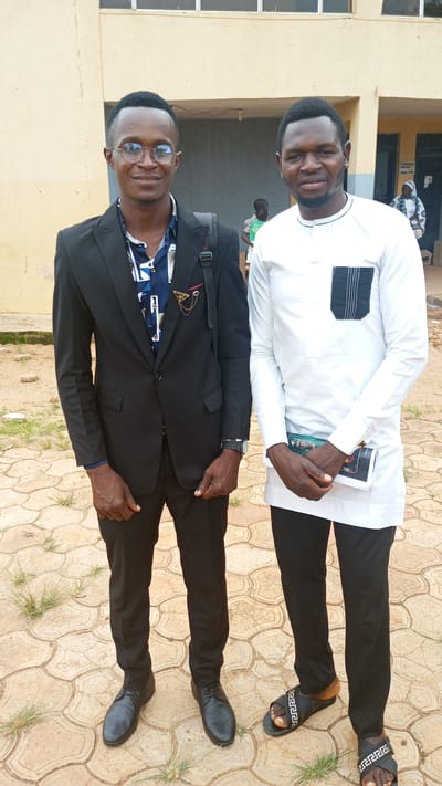 Two men stand side-by-side outdoors on a paved surface with beige interlocking geometric patterns, likely in Barikin Sabon Gidan Kanat, Nigeria. The man on the left wears a black suit with a patterned shirt, glasses, black shoes, and carries a black bag over his shoulder. He also wears a wristwatch. The man on the right wears a white long-sleeved shirt with a black-trimmed pocket, black pants, and sandals with a Greek key design. He holds a book or document in his hands. In the background, a tan building with visible doors and windows, and a few indistinct people, are partially visible. Grass grows sparsely between the paving stones.