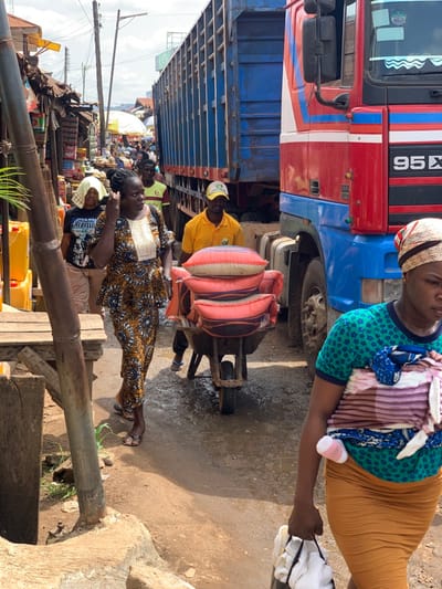 The image depicts a scene in Ondo, Nigeria (coordinates: 7.251, 5.196). A large, red and blue articulated lorry is parked on a narrow dirt road. A man in a yellow shirt is pushing a wheelbarrow full of sacks of rice past the truck. A woman in a brown and black dress is walking in the opposite direction. Another woman, wearing a green and white shirt, is walking toward the camera, carrying a white bag and a baby wrapped in a pink and white striped blanket. A woman in a beige shirt and blue jeans is walking behind the woman in the brown and black dress. The scene is set in a congested urban area, likely a marketplace, with buildings and shops lining the street. The ground is dirty and uneven, and the air is hazy with dust. There are power lines and poles running overhead. The image is captured in a low-angle perspective, focusing on the foreground and the interaction between the people. The scene is bustling with activity, reflecting the dynamism and energy of daily life in an urban area of Nigeria.