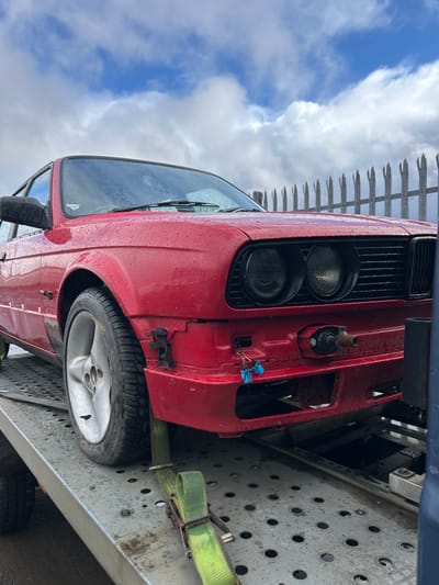 A red car with a missing front bumper is secured with a green strap on a gray metal trailer. The car's tire is visible on the trailer. The sky is partially cloudy. The location is Shotts, United Kingdom.