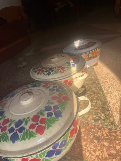 The scene, located within a residence in Isiamigbo, Nigeria, displays three lidded food warmers arranged diagonally on a tiled floor under direct sunlight. The foreground object is a large, off-white enamelware food warmer with a lid, adorned with a repeating pattern of red and blue flowers with green leaves, and equipped with two side handles. Immediately behind it is a smaller, matching off-white enamelware warmer with the identical floral pattern and two handles. In the background, a third, smallest food warmer is white and features a distinct multi-colored, possibly stylized figurative pattern around its circumference. All objects are illuminated by strong, direct sunlight, casting shadows on the floor. The floor is composed of patterned tiles, exhibiting sections of mottled green-brown and orange-brown hues. The upper left background, predominantly in shadow, reveals a partial view of dark red upholstered furniture and two light-colored shoes or sandals resting on the floor. No individuals are present, and no actions or interactions are depicted.