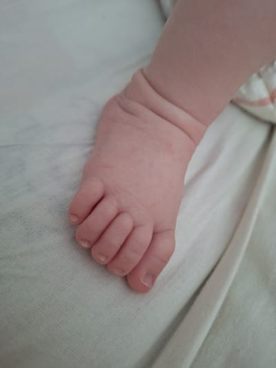 A close-up, high-angle shot focuses on the left foot of an infant, resting on a white, textured surface that appears to be bedding. The foot is positioned with the toes slightly curled and angled towards the right of the frame. The infant's skin is pale and smooth, with visible creases on the ankle and the base of the toes. The toenails are small and appear to be clear. The background is out of focus, but the white fabric continues across the image, with some subtle folds and shadows indicating the surrounding environment. The lighting is soft and even, suggesting an indoor setting. There is no visible indication of the city of Новобурейский, Russia, or any other specific location.