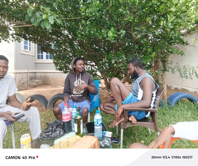 Three men are gathered outdoors, seemingly in a yard or garden area. The man on the left is wearing a striped white and blue shirt and striped pants. He is holding a small electronic device in his left hand and making a hand gesture. The man in the center is wearing a brown hooded sweatshirt and blue shorts. He appears to be talking or gesturing with his right hand. The man on the right is wearing a gray and white tank top and blue shorts. His legs are crossed, and he is looking down. A black garbage bag, several bottles of soda and water, a liquor bottle, and other items are on a glass table in front of them. In the background, there is a light-colored building with windows and a large tree with dense green foliage. Old car tires are scattered around the ground, and grass is growing. The image parameters, indicating it was taken with a CAMON 40 Pro camera, are displayed below the image.