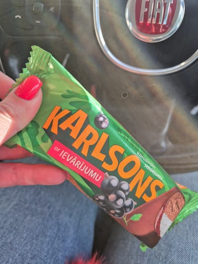 A hand with red nails holds a green Karlsons ar Ievarijumu black currant ice cream bar in front of a Fiat steering wheel and blue jeans. The image appears to be taken indoors, possibly inside the car. The expiration date on the bar is visible as 11.04.2023. The scene does not provide sufficient information to determine a specific location within the Breckland District or even the United Kingdom.