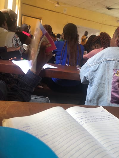 In a classroom setting, likely a university lecture hall, several young adults are seated at wooden desks, facing towards the front where a male lecturer, dressed in an orange shirt, is addressing them. The room has yellow-painted walls and a white ceiling with a ceiling fan. Sunlight streams in through tall, vertical windows on the left side of the image.
In the foreground, a notebook lies open on a desk. The pages contain handwritten notes in blue ink. Some of the visible text includes terms like "Variables," "Hypothesis," "Concept," and "Sampling." Another open notebook is visible further to the right, with more handwritten text.
Several students are visible, mostly from the back or side. One student in the immediate foreground is wearing a plaid shirt and is holding up a colorful book. Another student to their right wears a light blue denim jacket. Other students are wearing blue tops and pink tops. The general atmosphere appears to be one of focused learning, with students engaged in listening to the lecture and taking notes.
While the exact location of Achara Ndiagu Omegio, Nigeria is not directly discernible from the image, the scene depicts a typical educational environment within a community that could be located in Nigeria. The presence of a lecturer and students, along with the classroom setup, suggests an academic institution.
