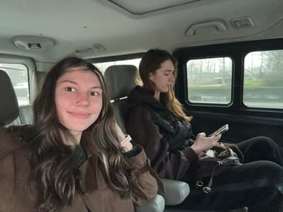 Two young adult females are depicted as passengers in the rear compartment of a vehicle in Huangdao District, China. The vehicle's interior features gray upholstered seating, headrests, and ceiling panels, contrasted with dark window frames.
The female positioned in the foreground, seated on the left, has long, wavy dark brown hair with lighter highlights. She wears a brown jacket with a suede-like texture over a dark green garment. A small stud piercing is visible on her left nostril, and a light-colored smartwatch is present on her left wrist. She is looking slightly to her right, displaying a subtle smile, with her right arm resting on the adjacent seat's armrest.
The second female, seated further back on the right, has long, straight reddish-brown hair. She is dressed in a dark brown long-sleeved top, a black quilted vest, and black pants. Her attention is focused downward on a silver-colored smartphone, which she holds with both hands. A dark brown handbag with gold-colored hardware rests on her lap.
Through the vehicle's windows, an overcast outdoor environment is visible, characterized by trees with sparse foliage, green ground cover, and distant light-colored structures, indicative of an urban or suburban setting. No direct interaction is observed between the two individuals.