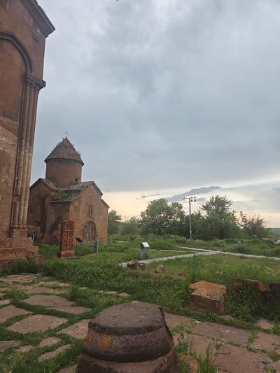 The image depicts a serene, overcast scene at a historical site in Vahramaberd, Armenia. In the foreground, a weathered stone column base, possibly part of an ancient structure, rests on a path paved with large, irregular stone slabs interspersed with grass. Beyond this, a small, old stone church with a conical roof and a cross at its apex stands prominently, its reddish-brown stone facade showing signs of age and vegetation growth. In front of the church, a carved stone monument, resembling a khachkar, is visible. The surrounding area is covered in lush green grass and scattered rocks, with trees and bushes forming a backdrop. A lamppost with multiple lights stands further in the distance, near a path. The sky is a mix of gray clouds and soft, diffused light, suggesting either early morning or late evening. There are no people or animals present in the image.