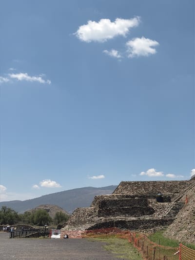 The image depicts the Teotihuacan pyramid in San Francisco Mazapa, Mexico, under a clear blue sky with scattered white clouds. A portion of the pyramid's stone structure is visible, featuring multiple levels and steps. An orange safety barrier runs alongside the pyramid. A mountain is visible in the background. On the ground, there's a paved pathway with sparse vegetation. In the distance, people are visible at the base of the pyramid.